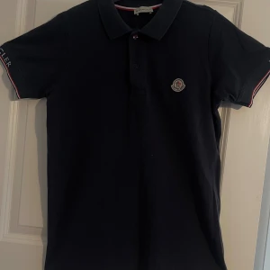 Svart pikétröja från Moncler - Säljer en snygg svart pikétröja från Moncler. Den har en klassisk krage och knappar framtill. Tröjan har korta ärmar med en liten Moncler-logga på bröstet. Perfekt för en stilren och avslappnad look!