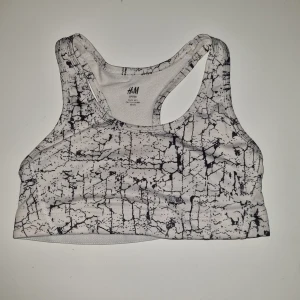 Vit och svart sport-bh från H&M - Säljer en cool vit och svart sport-bh från H&M med ett unikt sprucket mönster. Den är perfekt för träning och har en bekväm passform med brottarrygg. Materialet känns mjukt och stretchigt, vilket gör den idealisk för alla typer av träningspass. Är sprucken i sömmen,  lätt att fixa om man är händig.