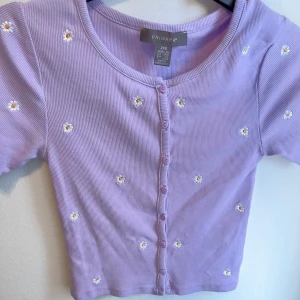 Lila ribbad topp med blommor från Primark - Säljer en superfin lila ribbad topp från Primark. Den har korta ärmar och är dekorerad med små söta blommor över hela tyget. Perfekt för vår och sommar! Toppen har knappar framtill och är i ett mjukt och bekvämt material.