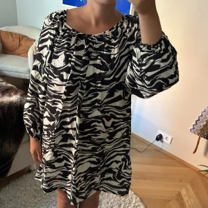 Svartvit mönstrad klänning från H&M - Säljer en superfin svartvit klänning från H&M med ett coolt zebramönster. Klänningen har långa ärmar och en lös passform som gör den perfekt för både vardag och fest. Materialet känns lätt och bekvämt, perfekt för vår och sommar!
