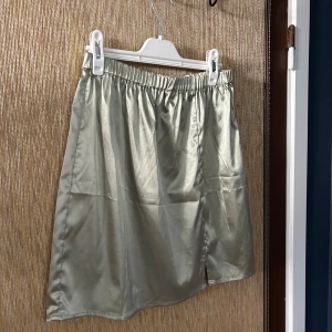 Silvrig kjol med resårmidja - Säljer en snygg ljus grön kjol med resårmidja. Kjolen är i ett glansigt material och har en liten slits på ena sidan. Perfekt för festliga tillfällen eller när du vill glittra lite extra! 🥳
