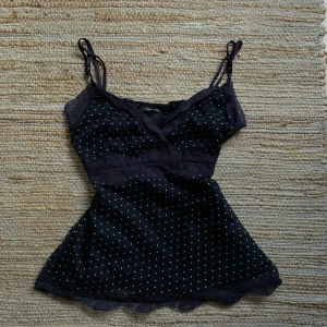 Svart prickig linne från Vero Moda - Säljer ett svart linne från Vero Moda med vita prickar. Det har spetsdetaljer och tunna axelband. Perfekt för en somrig look eller en festkväll!