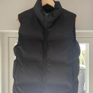 Svart dunväst från Knowledge Cotton Apparel - Säljer en snygg svart dunväst från Knowledge Cotton Apparel i storlek S. Perfekt för höst och vinter när du vill hålla dig varm men ändå se stilren ut. Västen är i nyskick och har en hög krage för extra värme. Nypris ligger på 1499 kr.