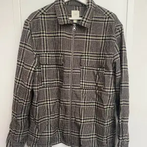 Säljer en snygg rutig overshirt från H&M i svart och vit. Den har en dragkedja framtill och långa ärmar. Perfekt för höst och vår, och funkar både till vardags och fest. Storleken är herr M. Passar bra över en t-shirt eller tröja för en avslappnad look. Skick: Mycket bra - Artnr: C051