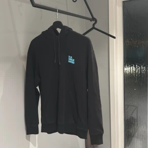 Svart hoodie från H&M med tryck - Säljer en svart hoodie från H&M i storlek S. Den har ett coolt tryck i turkos på baksidan och text på framsidan. Perfekt för en avslappnad stil med sin relaxed fit. Grym för höst och vinter!