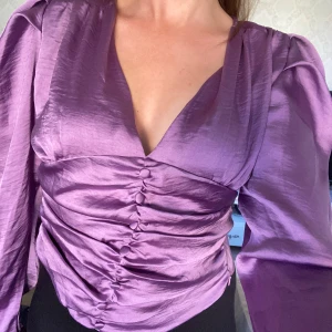 Lila satinblus från IVIVI - Säljer en superfin lila satinblus från IVIVI i storlek S. Blusen har en v-ringad design med knappar framtill och långa ärmar. Den har en smickrande rynkad detalj i midjan och elastisk rygg för en bekväm passform. Perfekt för både fest och vardag!