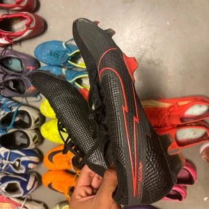 Svarta och röda fotbollsskor från Nike - Säljer ett par svarta Nike fotbollsskor med röda detaljer. Skorna har en snygg och modern design med snörning och är perfekta för fotbollsträning eller match. De har en slitstark sula med dobbar för bra grepp på planen. Använda men i bra skick!