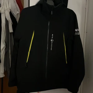 Svart jacka från Sail Racing - Säljer en svart jacka från Sail Racing i mycket bra skick. Den har en cool design med gula detaljer vid fickorna och en logga på ärmen. Perfekt för höst och vår, med en normal passform och långärmad stil. Jackan är gjord i ett slitstarkt material och har en praktisk huva.