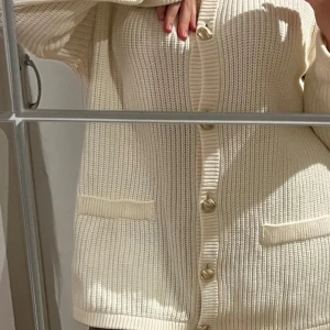 Vit stickad kofta - Säljer en elegant vit stickad kofta med guldiga knappar framtill. Perfekt för kyliga dagar, den har en avslappnad passform och två praktiska fickor. Koftan är långärmad och har en ribbad struktur som ger en klassisk look. Passar perfekt till både vardag och mysiga hemmakvällar. 