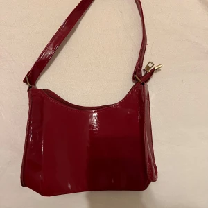 Röd axelväska i syntet - Snygg röd axelväska i glansigt syntetmaterial. Perfekt för en utekväll eller fest! Väskan har en praktisk dragkedja upptill och en stilren design som passar till många outfits. Den är i mycket bra skick och redo att användas direkt. Säljs för 40