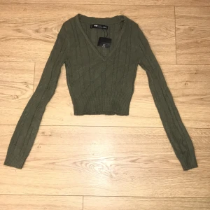 Olivgrön stickad tröja - Säljer en superfin olivgrön stickad tröja. Den är croppad med en snygg v-ringning och har långa ärmar. Perfekt för höst och vår när du vill vara både stilren och bekväm. Den är mycket stretch och är HELT oanvänd då den var en present. Tröjan är i mycket bra skick och redo för nya äventyr! 🍂💚