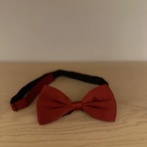 Röd fluga - Snygg röd fluga perfekt för festliga tillfällen. Den har en klassisk design med en justerbar rem i svart för bästa passform. Flugan är tillverkad i ett elegant material som ger en stilren look. Perfekt accessoar för att lyfta din outfit!