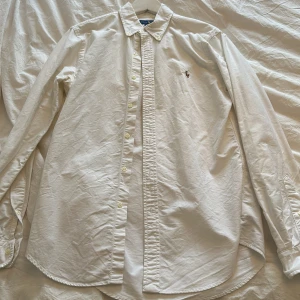 Vit oxfordskjorta Ralph lauren - Säljer en klassisk vit skjorta från Ralph Lauren i bomull. Den har en broderad logga på bröstet och knappar hela vägen ner. Perfekt för både vardag och fest! Skjortan är i bra skick och har långa ärmar. Storlek Medium slim fit