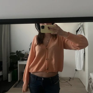 Persikofärgad kofta med knappar - Säljer en mysig persikofärgad kofta med knappar framtill. Koftan har en croppad passform och långa ärmar, perfekt för en avslappnad stil. Passar S/M