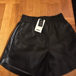 Svarta skinn shorts från Bik Bok - Snygga svarta shorts i syntetmaterial från Bik Bok. De har en elastisk midja för en bekväm passform. Perfekta för en stilren look. Prislappen finns även kvar! 