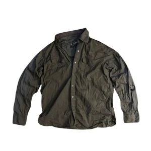 Grön workwearskjorta från Alpha Industries - Säljer en grön långärmad skjorta från Alpha Industries med broderade detaljer och knappar framtill. Skjortan har en cool militärinspirerad look och är perfekt för en avslappnad stil. Storlek XL. 