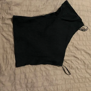 Svart oneshoulder topp - Snygg svart oneshoulder topp med ribbad textur. Perfekt för en stilren look. Den har en asymmetrisk design som ger en unik touch.