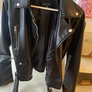 Svart skinnjacka från Amisu - Snygg svart skinnjacka från Amisu med dragkedja och knappar. Jackan har en klassisk bikerstil med en snedställd dragkedja och detaljer som en bröstficka med dragkedja och knappar på sidorna.