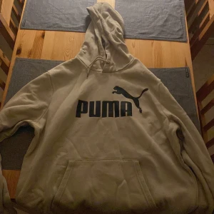 Beige hoodie från Puma - Säljer en beige hoodie från Puma med svart logga på bröstet. Den har en stor ficka framtill och en bekväm huva. Perfekt för en avslappnad stil.