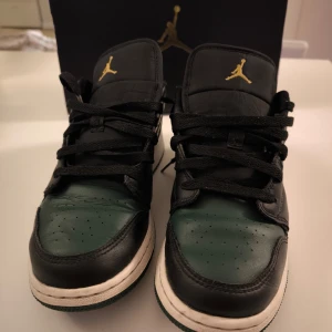 Nike Air Jordan 1 Low - Snygga Nike Air Jordan 1 Low sneakers i svart och grön med klassisk design. Skorna har snörning och en vit sula som kontrasterar mot det mörka lädret. Perfekta för en stilren look.