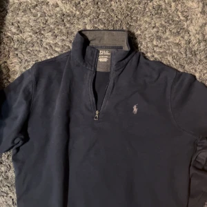 Mörkblå tröja från Ralph Lauren - Snygg mörkblå tröja från Ralph Lauren med halv dragkedja och klassisk krage. Perfekt för en stilren look. Den har en liten broderad logotyp på bröstet. Oanvänd