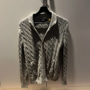 Grå stickad tröja med dragkedja från Polo Ralph Lauren - Säljer en stilren grå stickad tröja från Polo Ralph Lauren. Tröjan har en dragkedja framtill och ett diskret broderat märke på bröstet. Perfekt för kyliga dagar med sin kabelstickade design och långa ärmar.