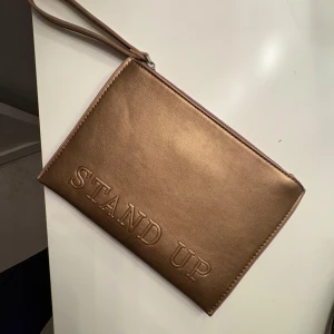 Bronsfärgad kuvertväska med text - Snygg bronsfärgad kuvertväska med texten 'STAND UP' präglad på framsidan. Väskan har en praktisk handledsrem och stängs med dragkedja. Perfekt för att bära med sig det viktigaste på ett stilfullt sätt.