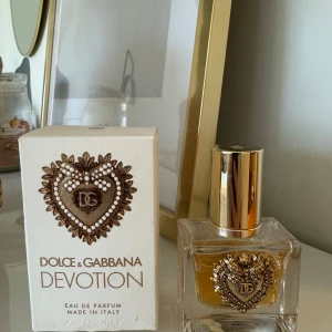 Dolce & Gabbana Devotion Eau de Parfum - Elegant parfym från Dolce & Gabbana med noter av kanderad citrus, apelsinblomma och vanilj. Flaskan har en lyxig design med ett dekorativt hjärta i guld på framsidan. Perfekt för den som älskar en sofistikerad och varm doft.