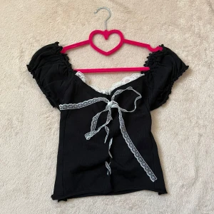 Svart topp med spetsdetaljer - Söt svart topp med korta puffärmar och vacker vit spets längs halsringningen. Toppen har en dekorativ snörning framtill med spetsband. 