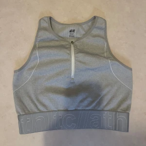 Grå sporttopp från H&M - Snygg grå sporttopp från H&M med dragkedja framtill. Perfekt för träning med ribbad design och bred resårkant med textdetaljer. Bekväm och stilren för alla dina sportaktiviteter.