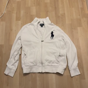 Vit tröja från Polo Ralph Lauren - Säljer en stilren vit jacka från Polo Ralph Lauren med dragkedja och det klassiska logotypbroderiet i mörkblått på bröstet. Jackan är tillverkad i bomull och har långa ärmar samt ribbade muddar.