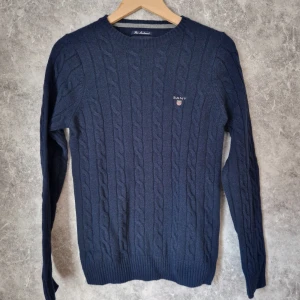 Mörkblå kabelstickad tröja från Gant lambswool - Snygg mörkblå kabelstickad tröja från Gant med broderad logga på bröstet. Perfekt för kyliga dagar med sin klassiska design och långa ärmar. Ribbstickade muddar ger en skön passform. 100% fine lambswool.