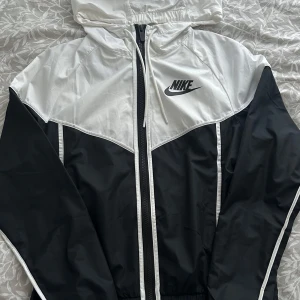 Svart och vit vindjacka från Nike - Snygg svart och vit vindjacka från Nike med dragkedja och huva. Jackan har ett klassiskt Nike-märke på bröstet och är perfekt för blåsiga dagar. Den är lätt och bekväm att bära. Oanvänd