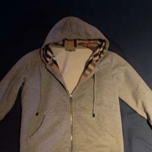 Grå huvtröja från Burberry - Säljer en stilren grå huvtröja från Burberry med dragkedja och klassiskt rutigt mönster på insidan av huvan. Perfekt för en avslappnad och trendig look. Tröjan är i strl s!