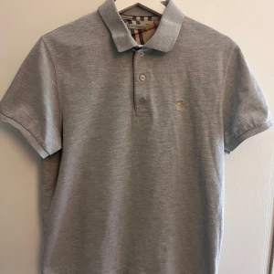 Grå pikétröja från Burberry - Säljer en stilren grå pikétröja från Burberry med klassisk krage och knappslå. Tröjan har ett diskret broderat emblem på bröstet och ikoniskt rutigt mönster på insidan av kragen. Perfekt för en avslappnad men elegant look.