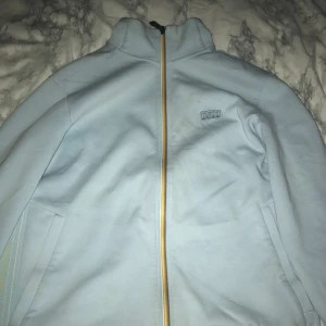 Ljusblå jacka från Boss - Snygg ljusblå jacka från Boss med dragkedja i kontrasterande gul färg. Jackan har en enkel design med två sidofickor och en diskret logga på bröstet. Perfekt för en stilren look.