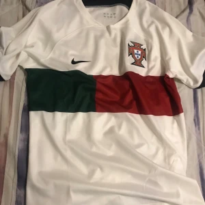 Portugal fotbollströja - Snygg vit fotbollströja med Portugals emblem på bröstet. Tröjan har en horisontell röd och grön rand över bröstet och korta ärmar. Säljer den eftersom den var för stor för mig, Storlek L