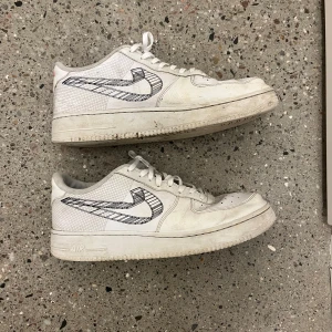 Vita Nike Air Force sneakers - Säljer ett par unika vita Nike Air Force sneakers med svart rutigt mönster på swoosh-loggan. Skorna har snörning och en bekväm sula med Air-dämpning. Perfekta för en stilren look.