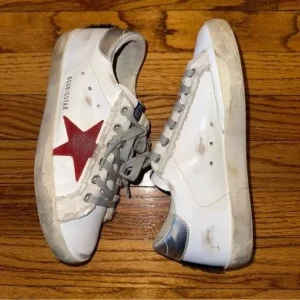 Vita sneakers från golden goose - Snygga vita sneakers från Golden Goose med en iögonfallande röd stjärna på sidan. Skorna har grå snörning och en silverdetalj vid hälen. Perfekta för en avslappnad stil med en touch av färg.