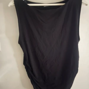 Svart ärmlös blus från Gina Tricot - Snygg svart ärmlös blus från Gina Tricot med en elegant rynkad detalj på sidan. Perfekt för en stilren look. Blusen har en enkel och tidlös design.