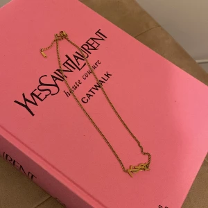 Guldhalsband med YSL-logga - Elegant guldhalsband från Yves Saint Laurent med den ikoniska YSL-loggan som hänge. Perfekt för att ge en touch av lyx till din outfit. Kedjan är tunn och stilren, vilket gör den lätt att bära till olika tillfällen.