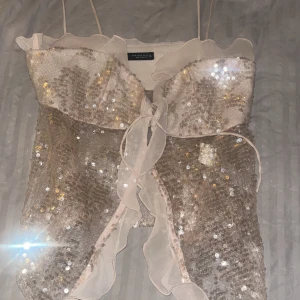 Glittrig paljettopp med volangdetaljer - Säljer en glittrig topp från zara med paljetter och volangdetaljer. Toppen har tunna axelband och en knytning framtill. Perfekt fest topp! Priset går att diskuteras 