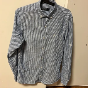 Randig skjorta från Polo Ralph Lauren - Snygg blå- och vitrandig skjorta från Polo Ralph Lauren med klassisk krage och knappar framtill. Skjortan har långa ärmar och en liten broderad logotyp på bröstet. Perfekt för en stilren look.