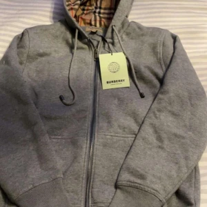 Burberry hoodie  - Säljer en stilren grå huvtröja från Burberry med dragkedja och klassiskt rutigt foder. Tröjan har en justerbar huva och långa ärmar, perfekt för en avslappnad look.