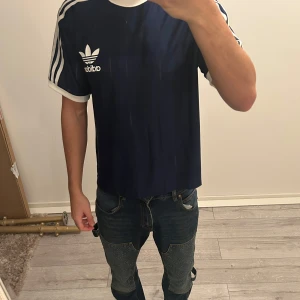 Blå t-shirt från Adidas - Snygg blå t-shirt från Adidas med klassiska vita ränder på ärmarna och logga på bröstet. Perfekt för en sportig look eller avslappnad stil. Kortärmad och bekväm passform.