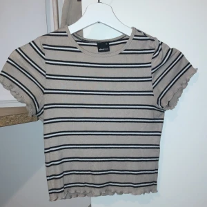 Randig ribbad topp från Gina Tricot - Säljer en beige och svart randig ribbad topp från Gina Tricot i storlek XS. Toppen har korta ärmar och en söt volangdetalj vid ärmslut och nederkant. Perfekt för en avslappnad stil. Säljer för att den inte är riktigt min stil längre plus att den är för liten. 