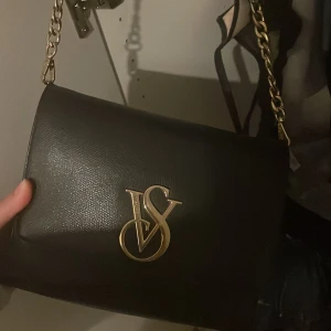 Svart axelväska med gulddetaljer - Elegant svart axelväska i skinn med en kedja i guld och ett stort VS-emblem på framsidan. Perfekt för att ge en lyxig touch till din outfit.