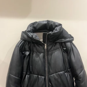 Svart pufferjacka från Zara - Snygg svart pufferjacka från Zara med dragkedja och hög krage. Jackan har en quiltad design och är perfekt för kyligare dagar. Den har en modern look och är tillverkad i syntetmaterial.