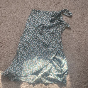 Blommig omlottkjol från Lindex - Säljer en fin blommig omlottkjol från Lindex i storlek XS. Kjolen har ett vackert mönster i grönt och vitt med blåa inslag. Perfekt för varmare dagar! en knapp är lite trasig men går fortfarande att använda som vanligt