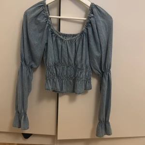 Blå jeansblus med puffärmar - Charmig blå jeansblus med puffiga långärmar och elastiska detaljer vid hals och midja. Perfekt för en avslappnad och stilfull look.från Gina 